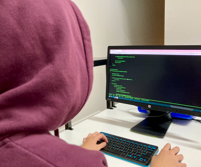 A girl with a hood on at a computer with her back to us fake coding